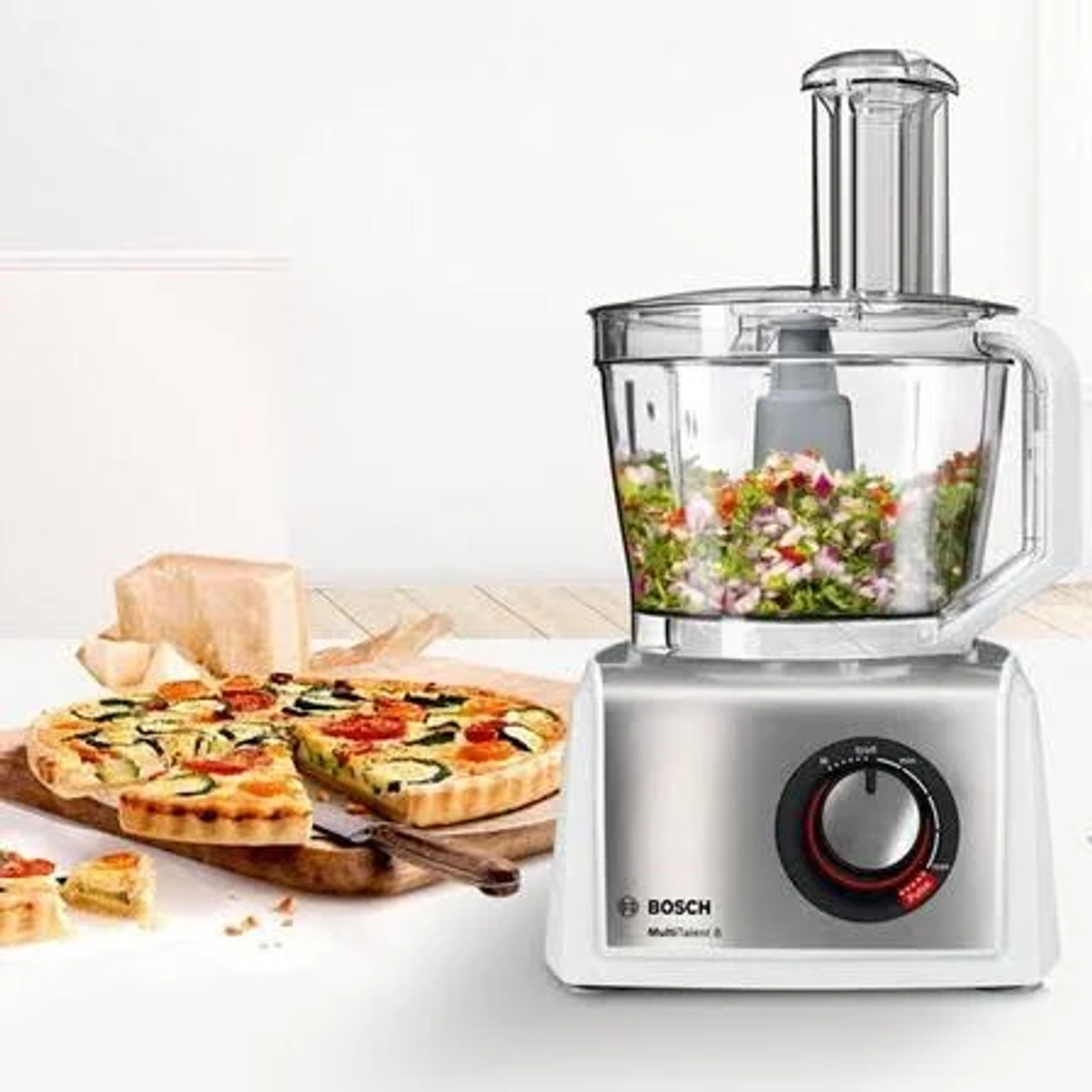 Un procesador de alimentos con una pizza e ingredientes. El procesador tiene verduras.