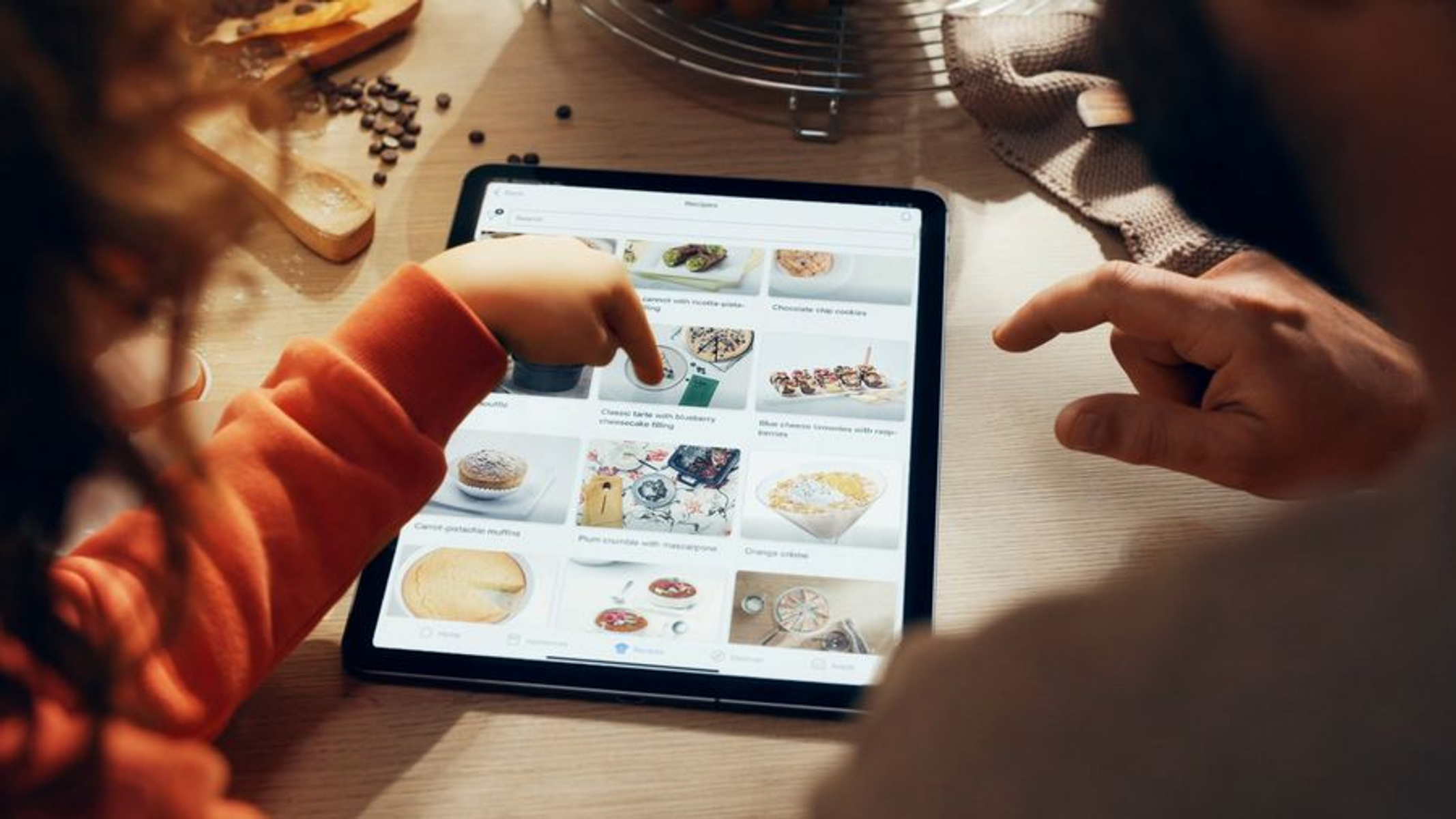 Dwie osoby przeglądają tablet, wybierając przepisy kulinarne. Ekran pokazuje zdjęcia potraw.