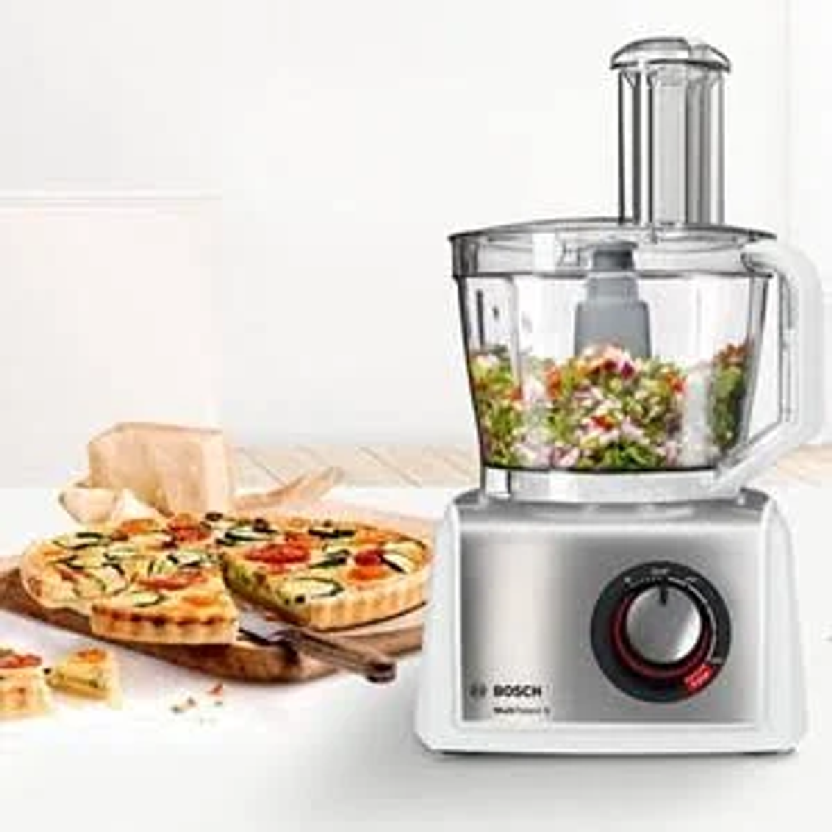 Procesador de alimentos con verduras picadas dentro, pizza e ingredientes sobre una mesa.