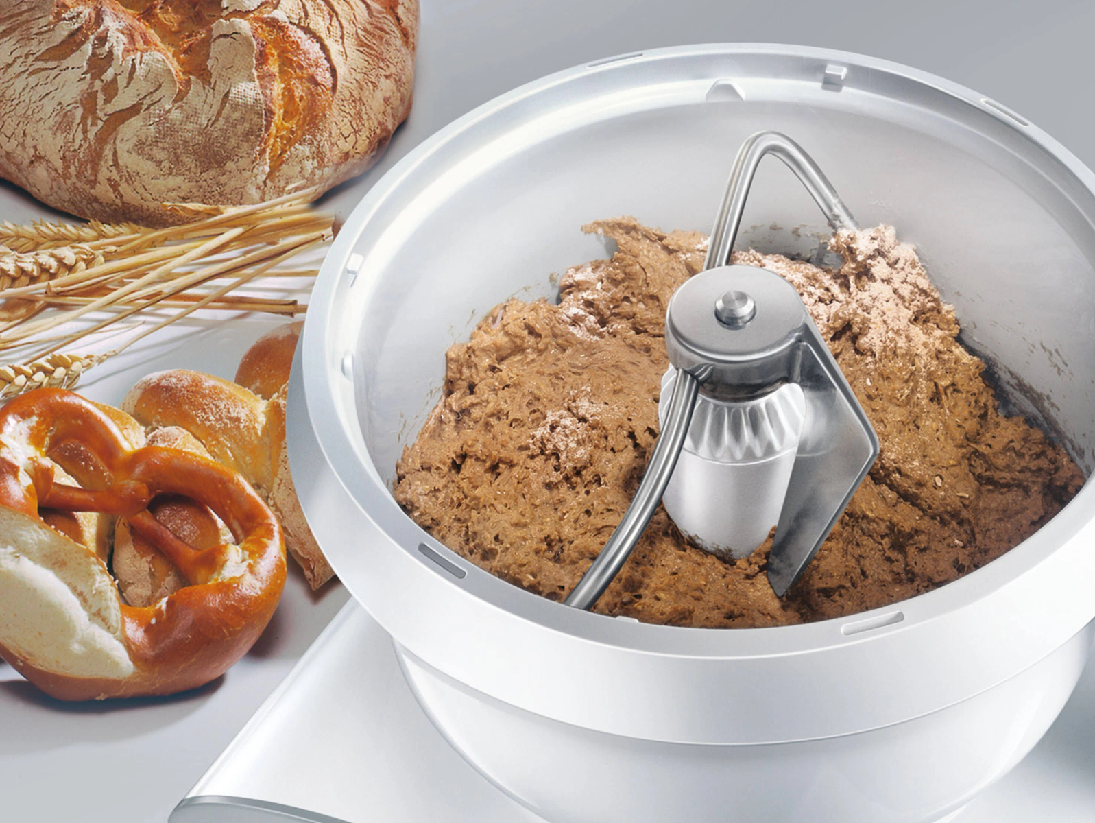 Een witte keukenmixer met deeg erin, met brood en een pretzel.