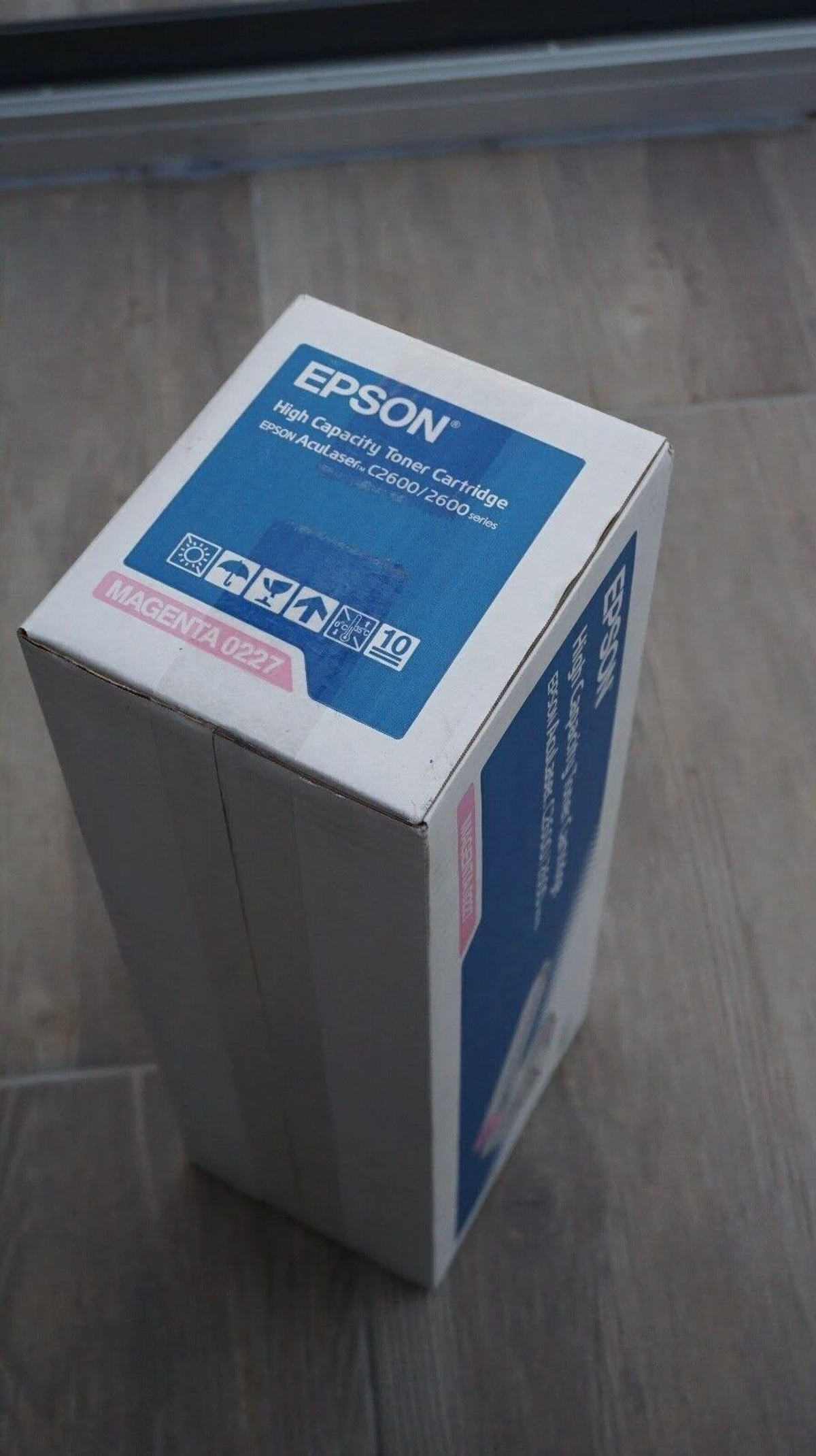 Una scatola di cartucce per stampanti Epson bianca e blu, angolata diagonalmente su un pavimento in legno.
