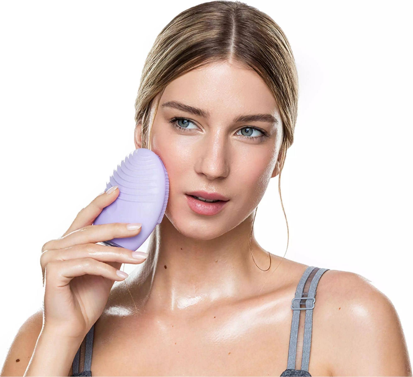 Una mujer de piel clara usa un cepillo de limpieza facial morado en su rostro.
