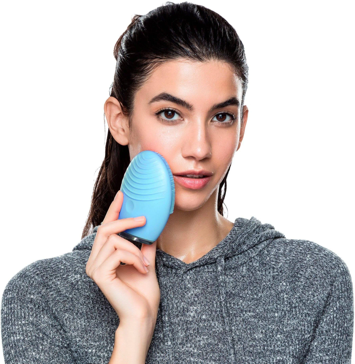 Mujer sosteniendo un dispositivo de limpieza facial azul en su mejilla. Lleva una sudadera gris.