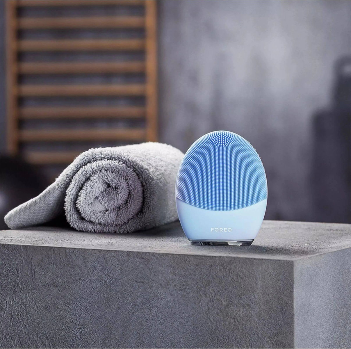 Limpiador facial Foreo azul y blanco sobre superficie gris, con toalla gris.