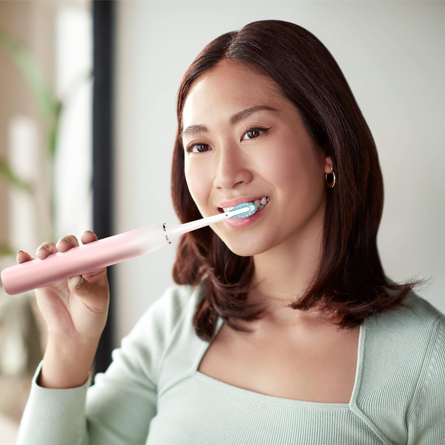 Una donna si lava i denti con uno spazzolino elettrico rosa. È in una stanza ben illuminata, sorridendo leggermente.