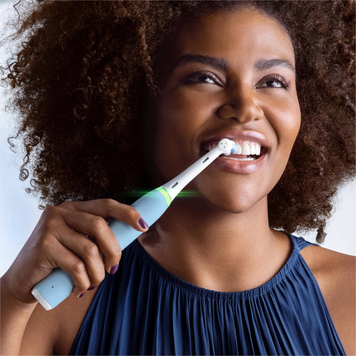 Mujer cepillándose los dientes con un cepillo de dientes eléctrico azul. Lleva una blusa azul oscuro.