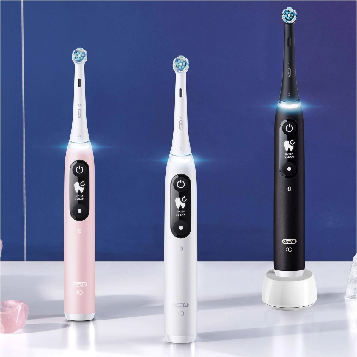 Three electric toothbrushes: pink, white, and black. Each has a brush head and a charging base.