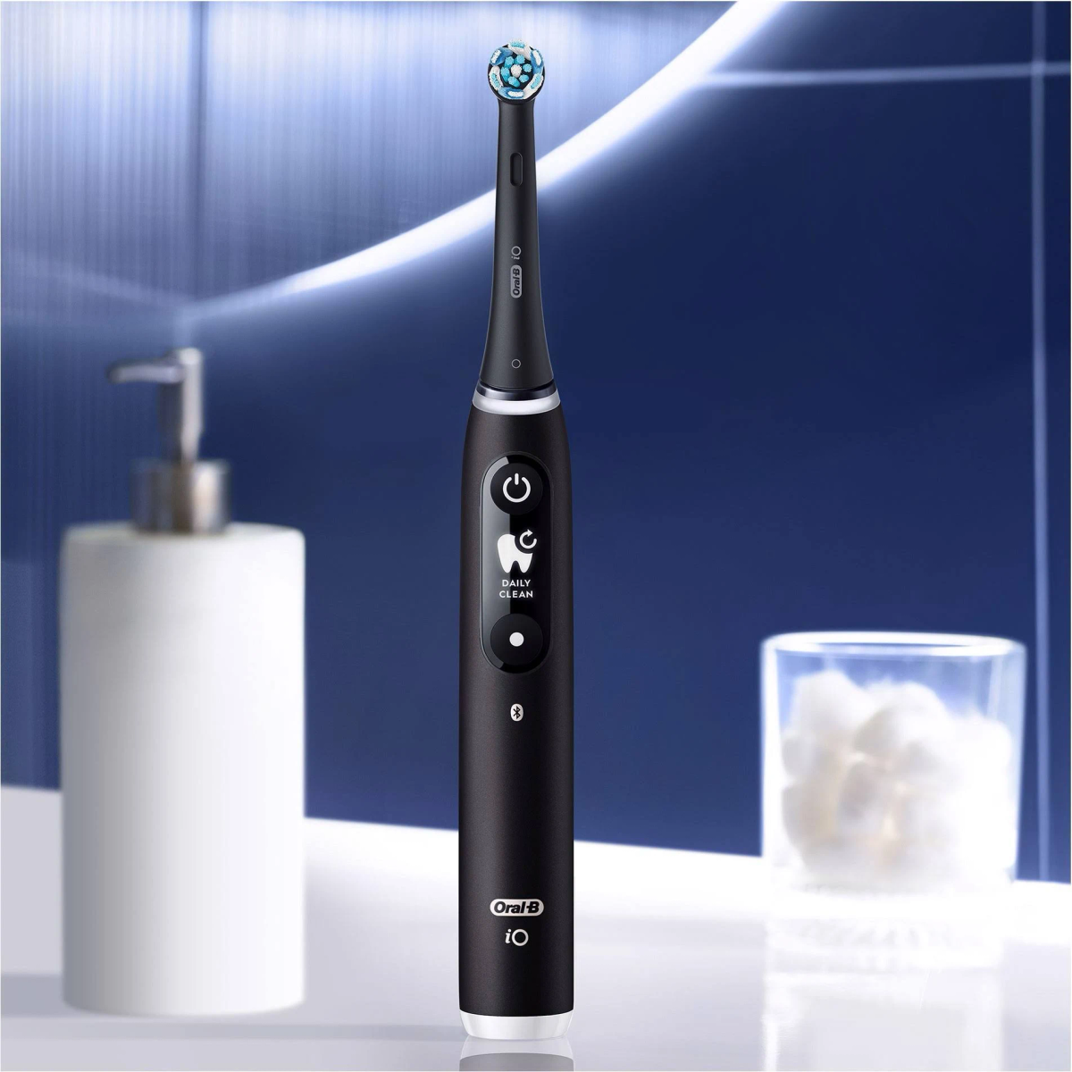 Black electric toothbrush with blue bristles, on a white surface, with a white soap dispenser and a glass with cotton.