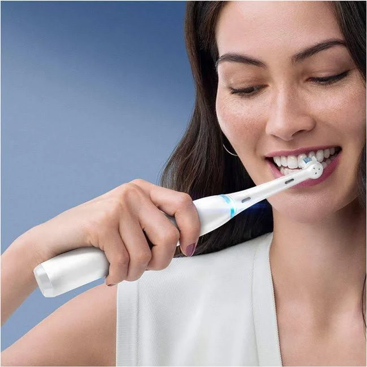 Mujer cepillándose los dientes con un cepillo eléctrico. Sonríe. El fondo es azul.