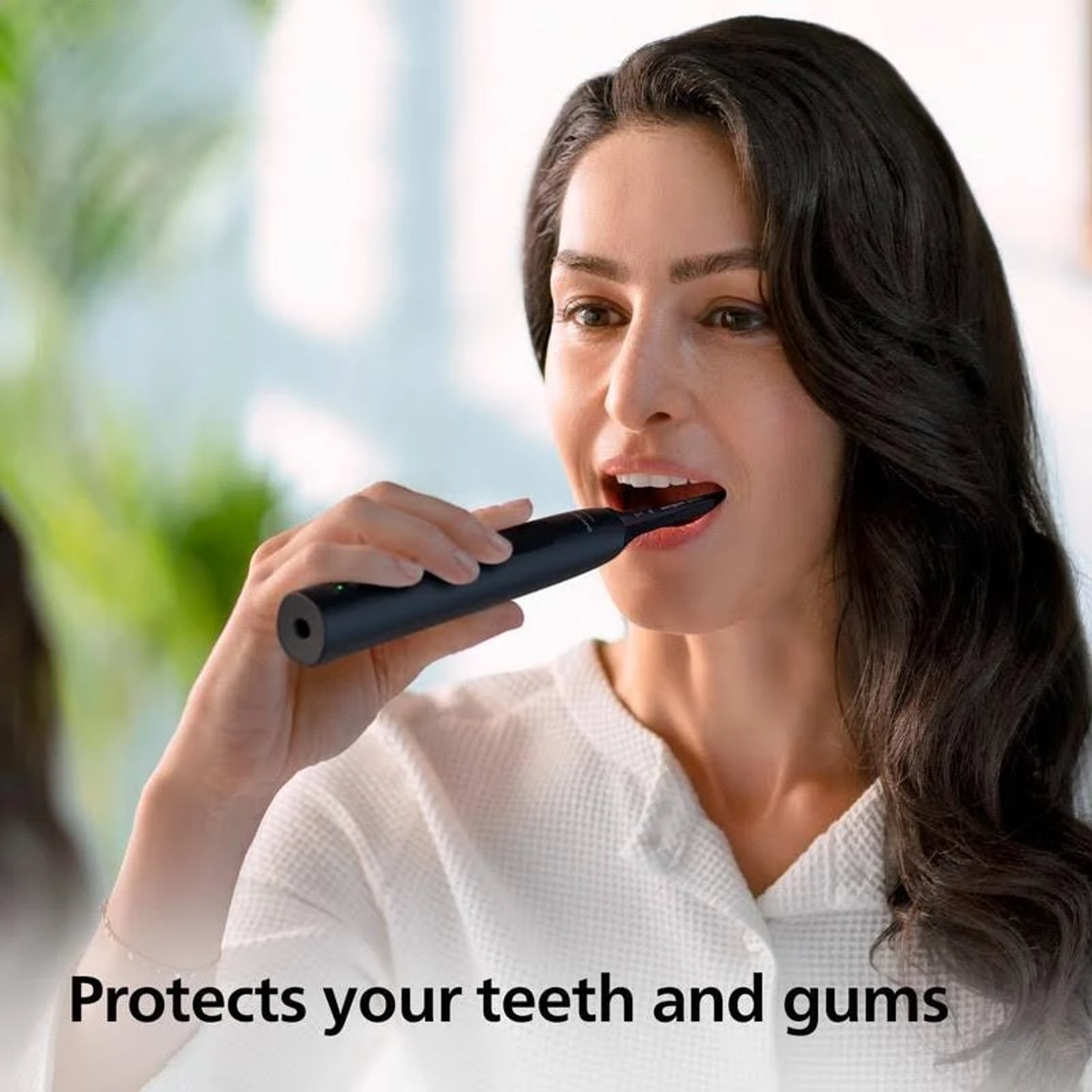 Kobieta myje zęby czarną elektryczną szczoteczką. Biały szlafrok, zielone tło.