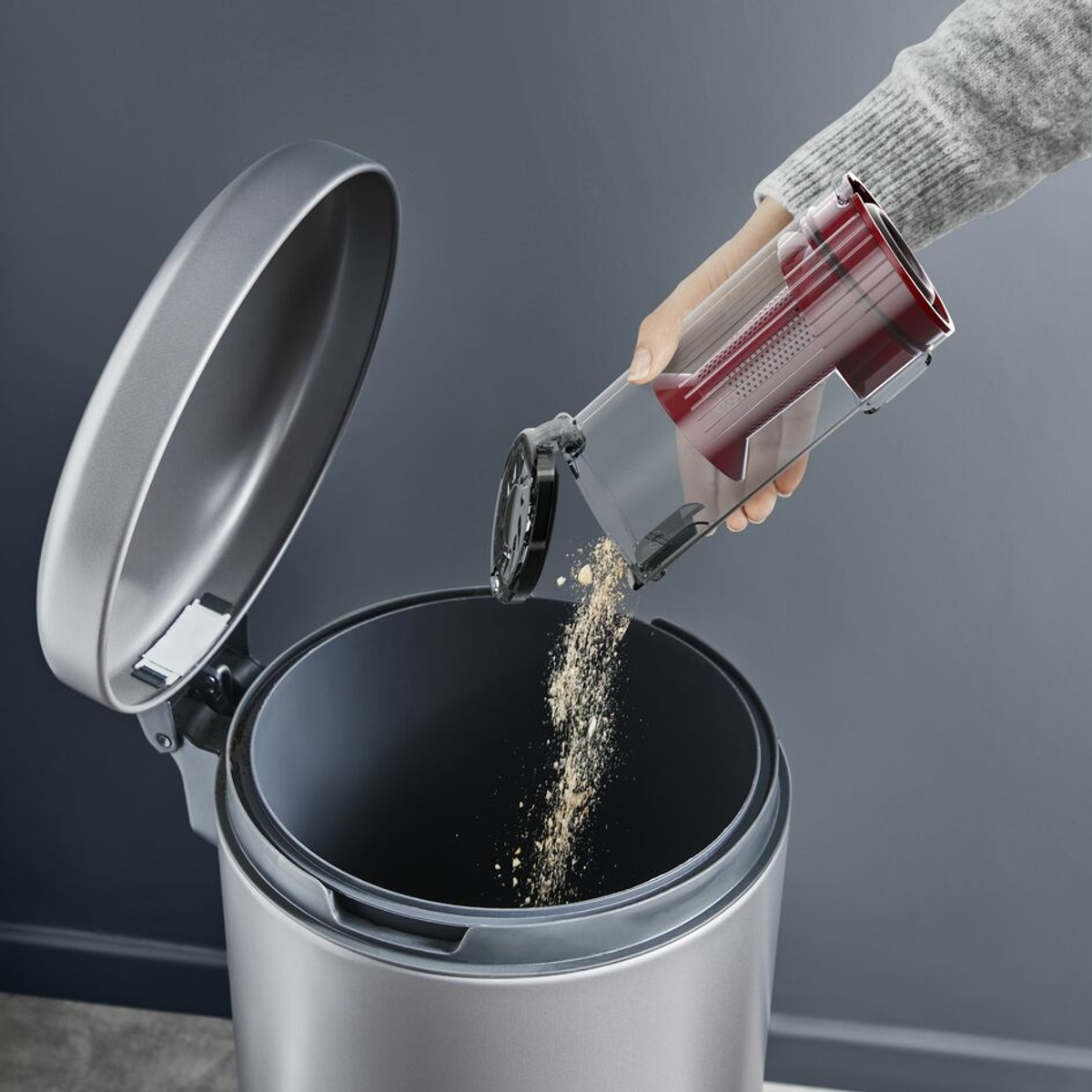 Una persona vaciando el depósito de polvo de una aspiradora en un cubo de basura. Fondo gris.