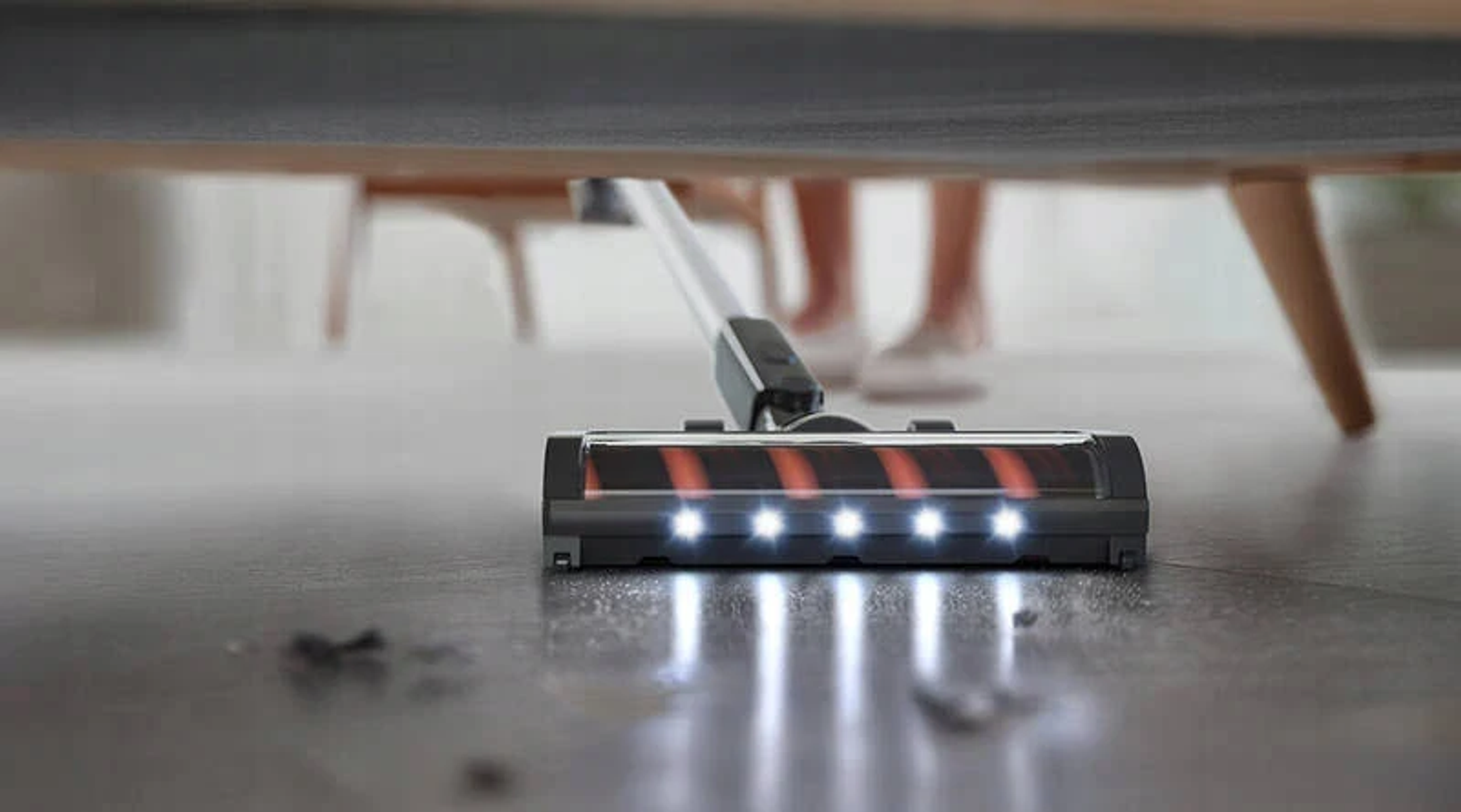 Una testina per aspirapolvere con luci sotto i mobili. Il pavimento è visibile. Piedi sullo sfondo.