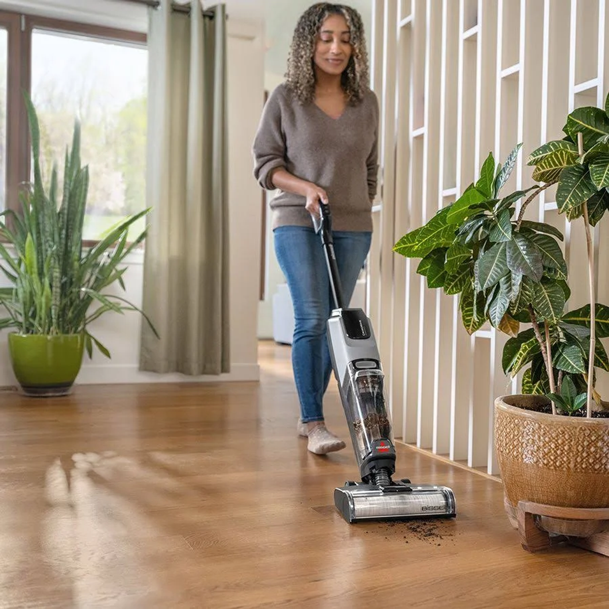 Una mujer aspirando un piso de madera con una aspiradora inalámbrica, plantas en el fondo.