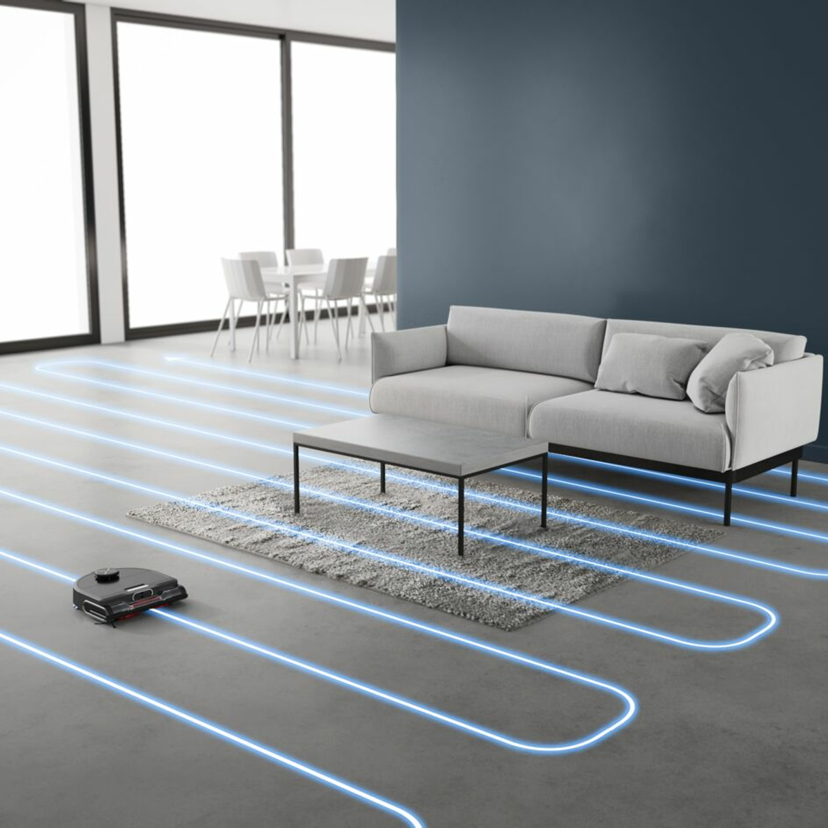 Robot aspirapolvere che pulisce una stanza. Divano grigio e tappeto sul pavimento. Planimetria visibile.