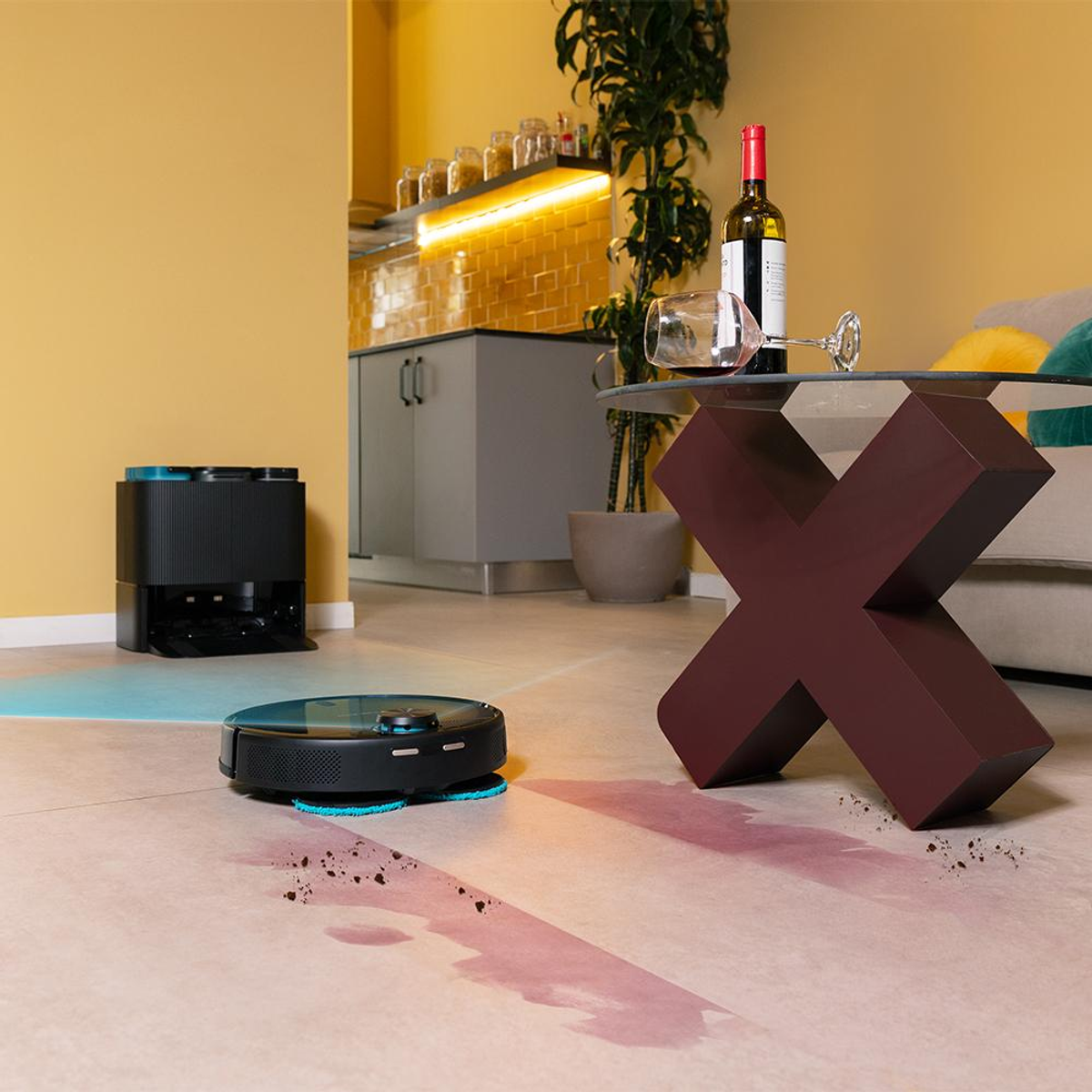 Un robot aspirapolvere su un pavimento piastrellato, con vino versato e un tavolo decorativo con una bottiglia di vino.