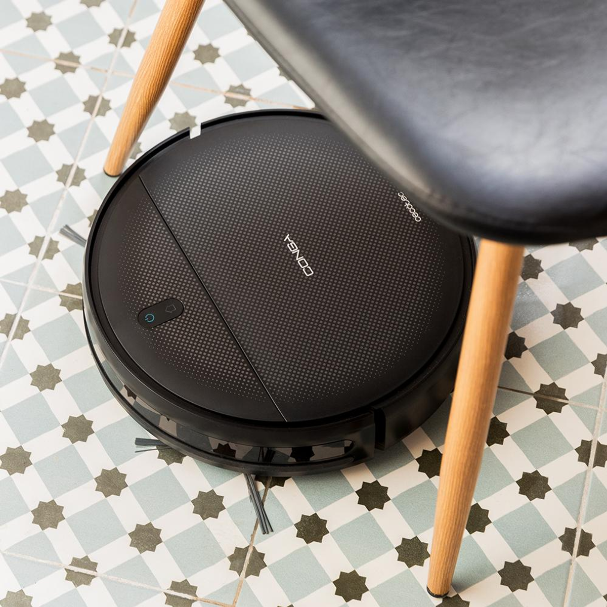 Czarny robot sprzątający pod krzesłem. Podłoga jest wyłożona płytkami z geometrycznym wzorem.