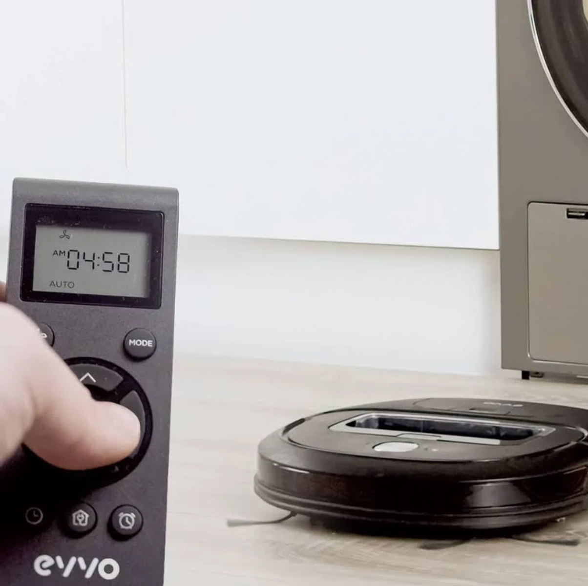 Mano che tiene un telecomando vicino a un robot aspirapolvere su un pavimento in legno.