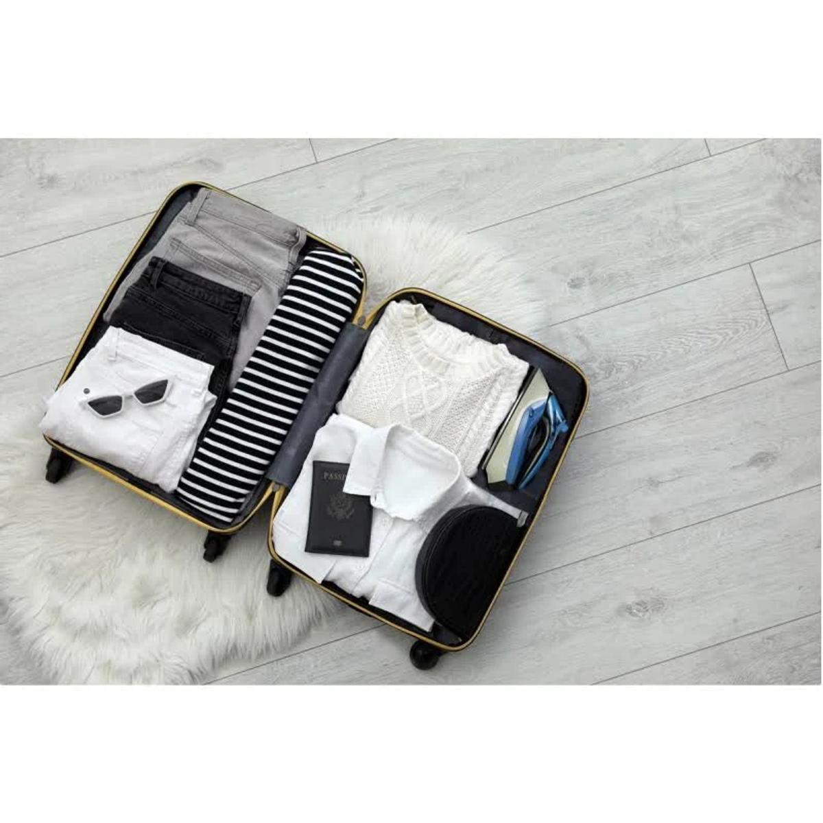 Otwarta walizka z ubraniami: dżinsy, sweter, biała koszula, akcesoria i paszport na dywanie.