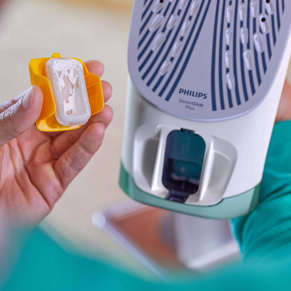 Ręka trzyma żółty pojemnik z białym prostokątnym przedmiotem obok białego i niebieskiego żelazka.