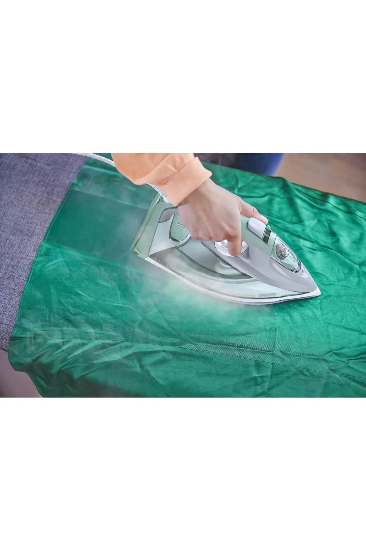 Dłoń prasuje zieloną tkaninę białym i szarym żelazkiem, widoczna para. Tło jest rozmyte.
