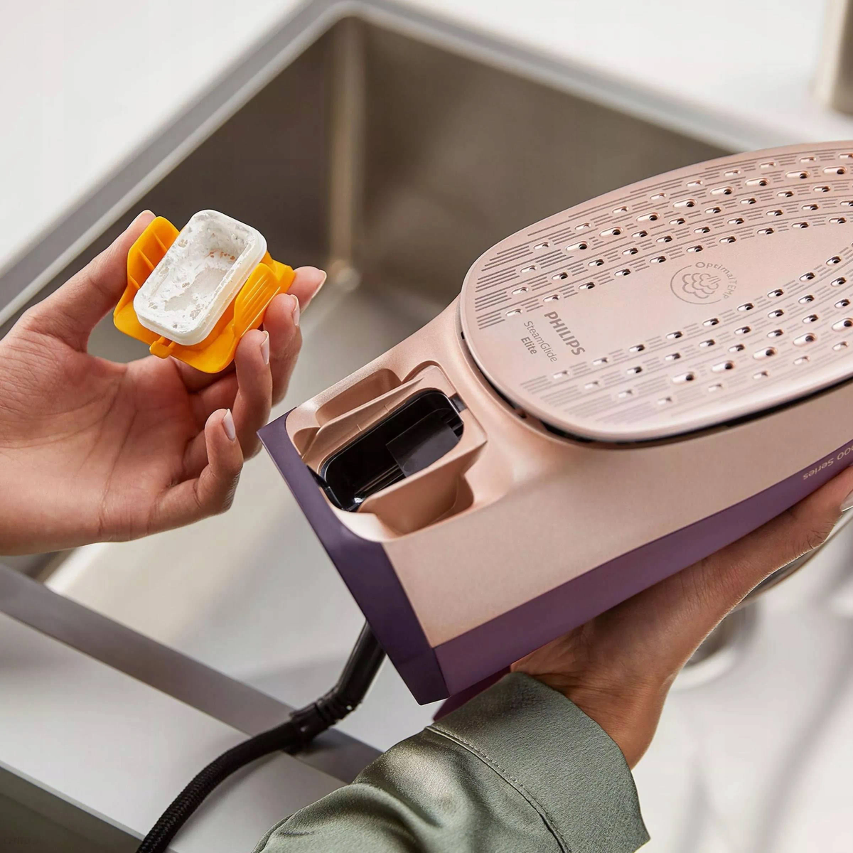 Dłonie trzymające żelazko Philips i zbiornik na wodę, z białym zlewem w tle.