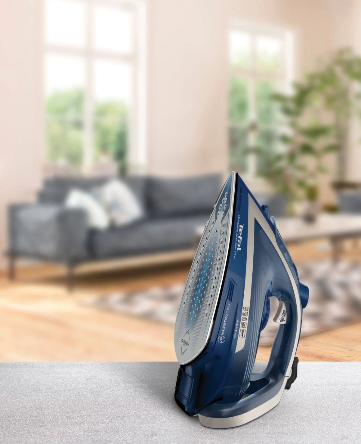Ein blaues und silbernes Bügeleisen auf einem Tisch, mit verschwommenem Wohnzimmerhintergrund. Das Bügeleisen ist angewinkelt.