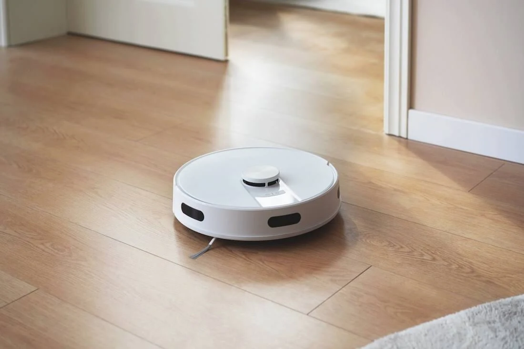 Biały, okrągły odkurzacz robot na drewnianej podłodze, blisko otwartych drzwi.
