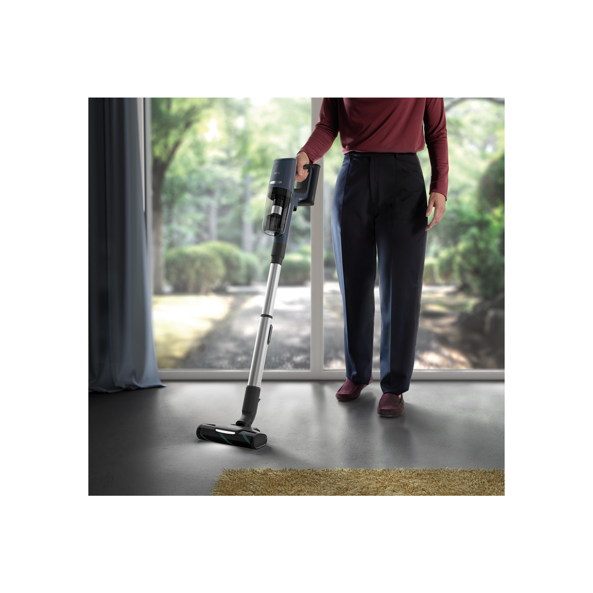 Persona aspirando con una aspiradora inalámbrica, en una habitación con ventana y alfombra.