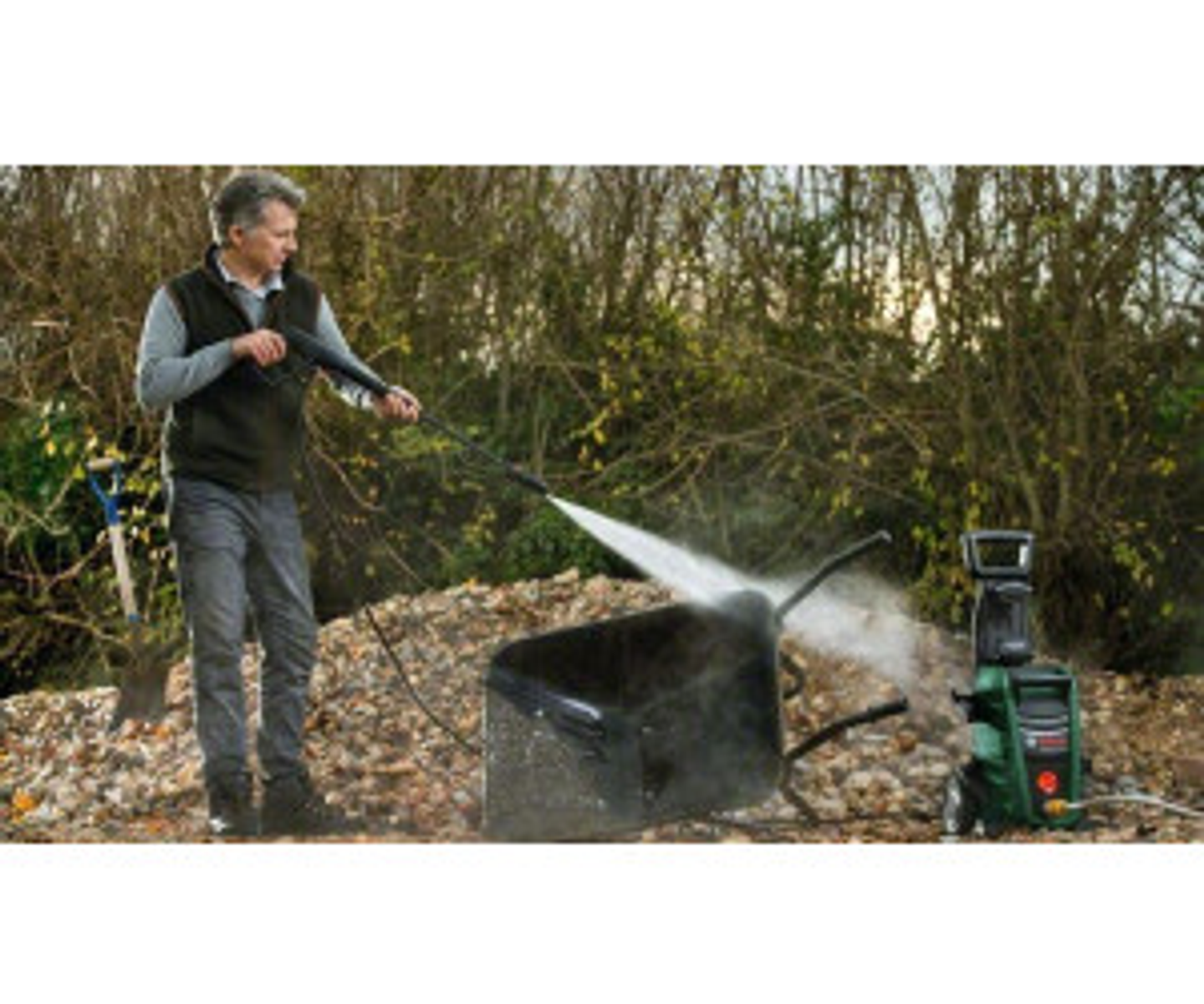 Homme pulvérisant de l'eau sur une brouette avec un nettoyeur haute pression, en extérieur.