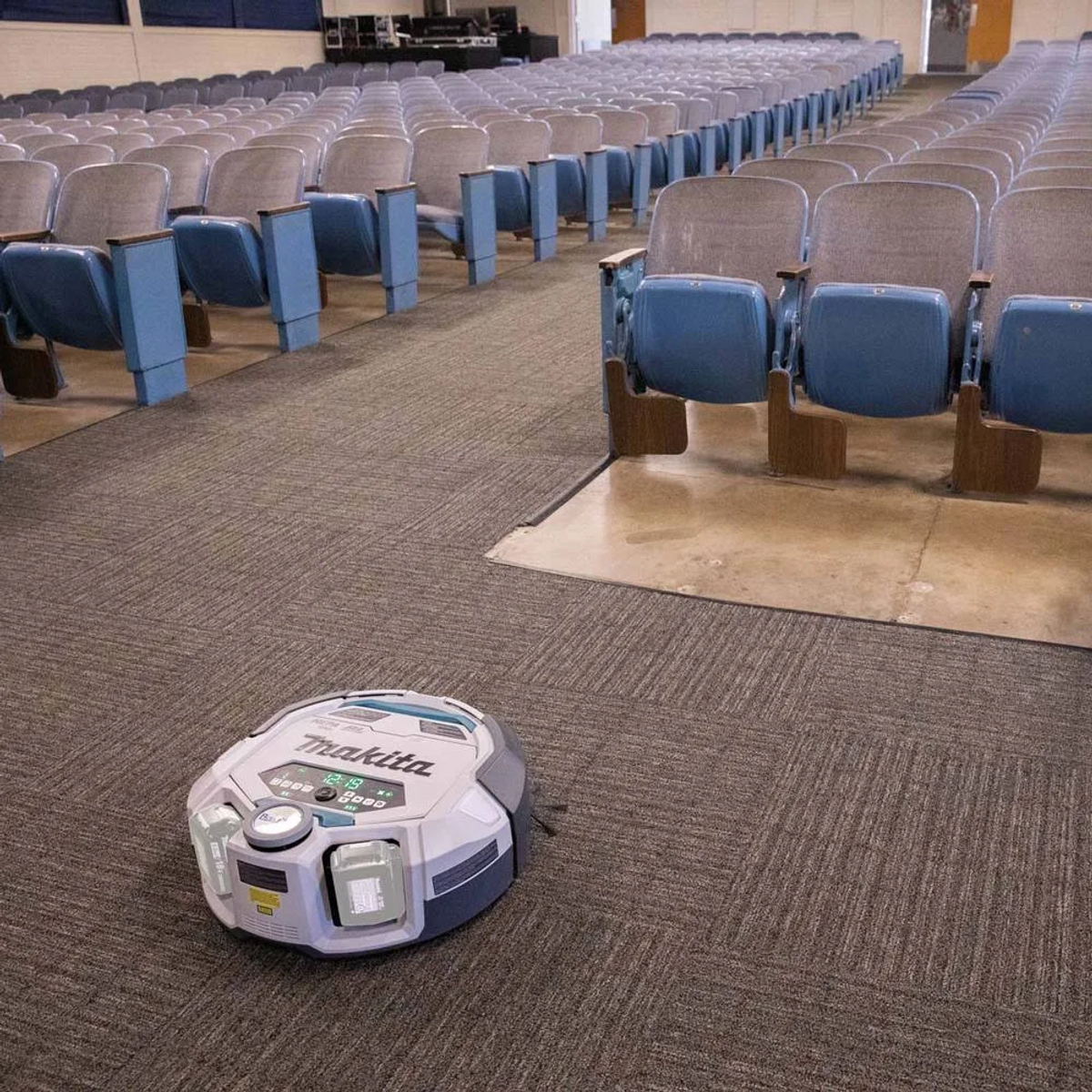 Aspirapolvere robotico su un pavimento in moquette in un auditorium con file di sedili blu.