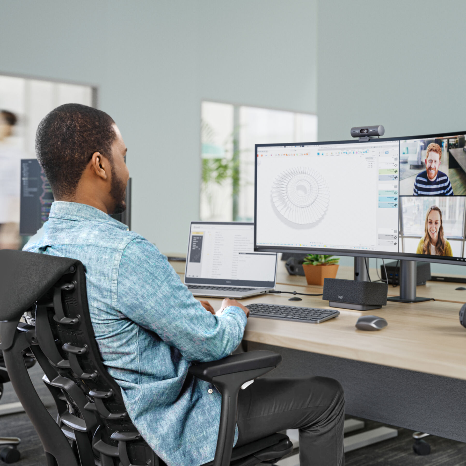 Mężczyzna w niebieskiej koszuli pracuje na komputerze, wideokonferencja na monitorach, otoczenie biurowe.