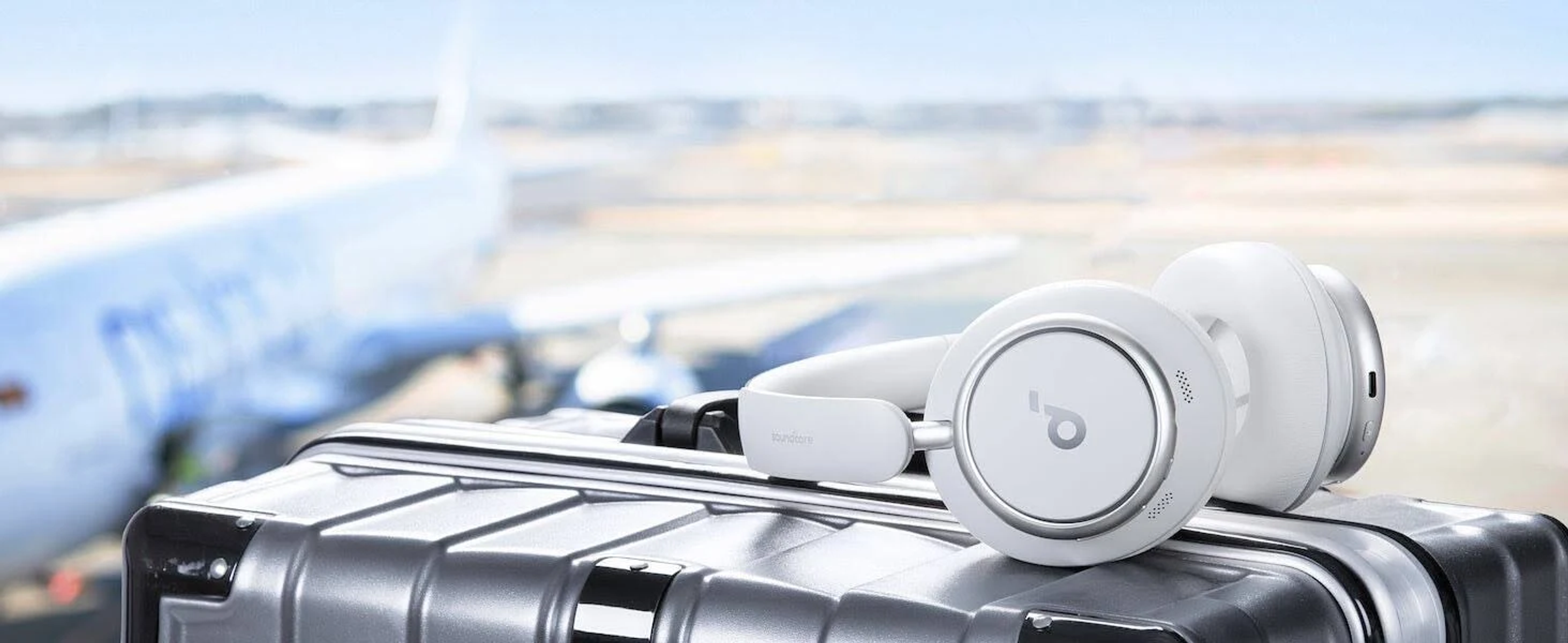 Białe słuchawki na srebrnej walizce, z samolotem w rozmytym tle.