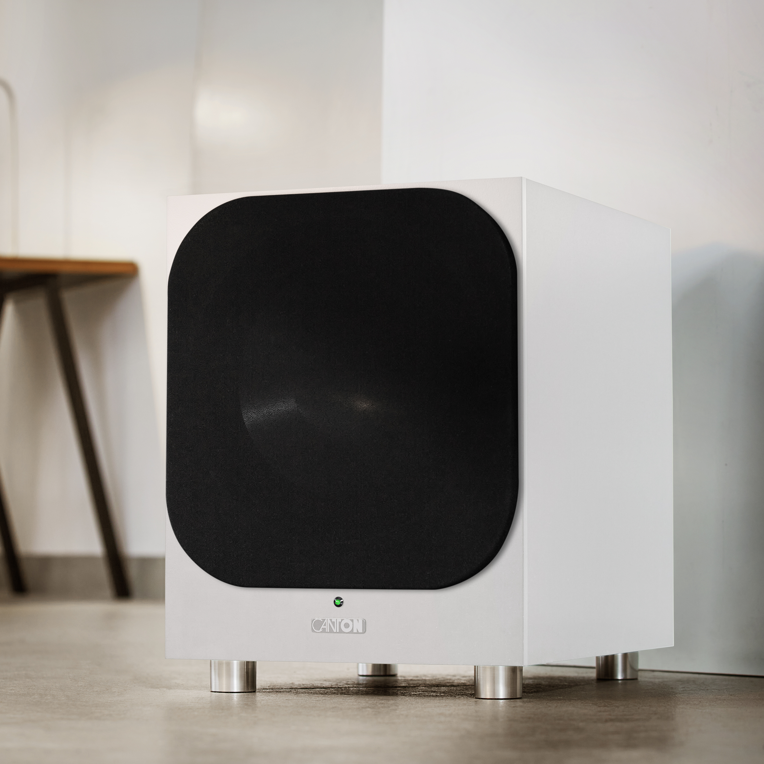 Weißer Subwoofer mit schwarzer Lautsprecherabdeckung und silbernen Beinen, in einem Raum.