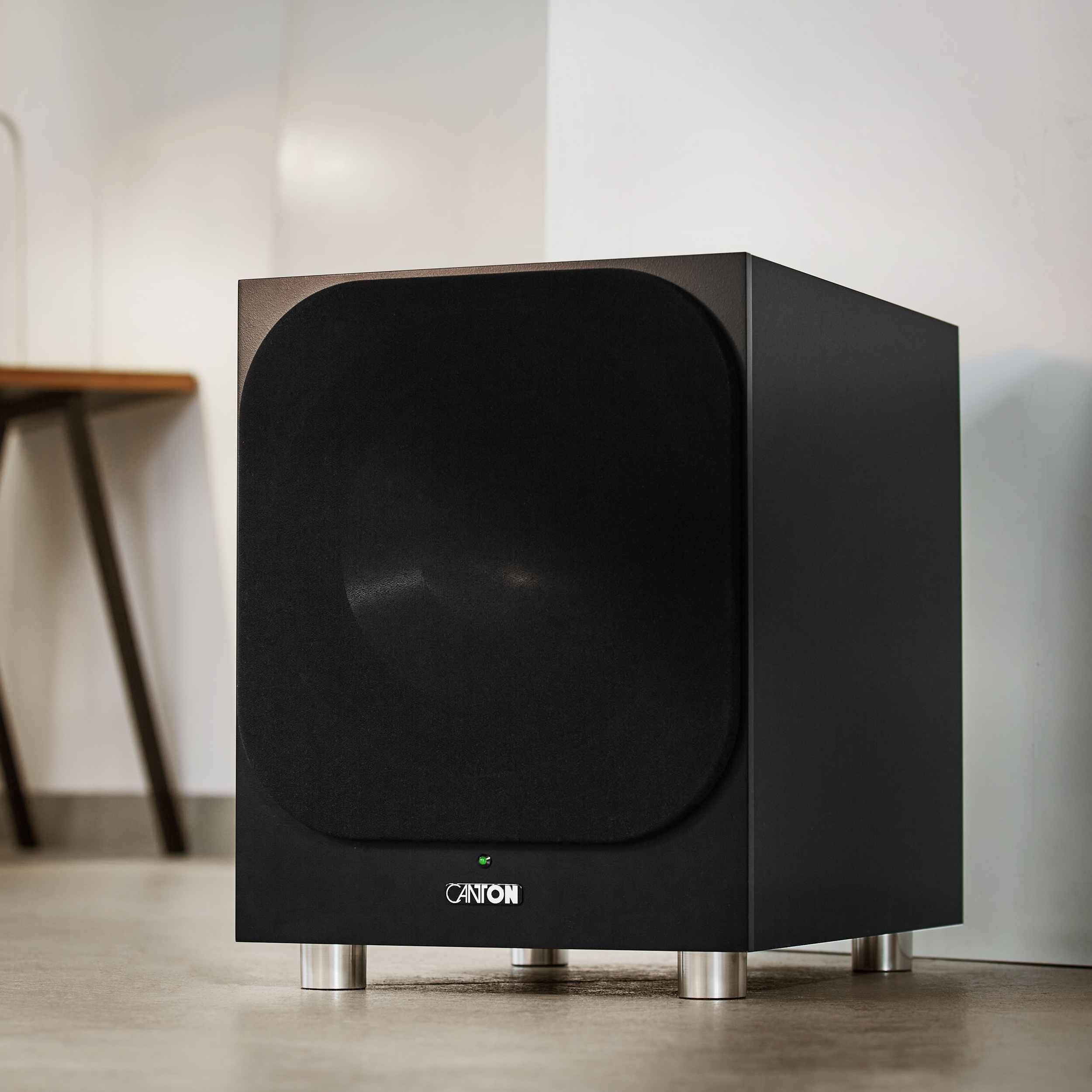 Schwarzer Subwoofer-Lautsprecher mit silbernen Beinen. Er steht auf einem Holzboden an einer weißen Wand.
