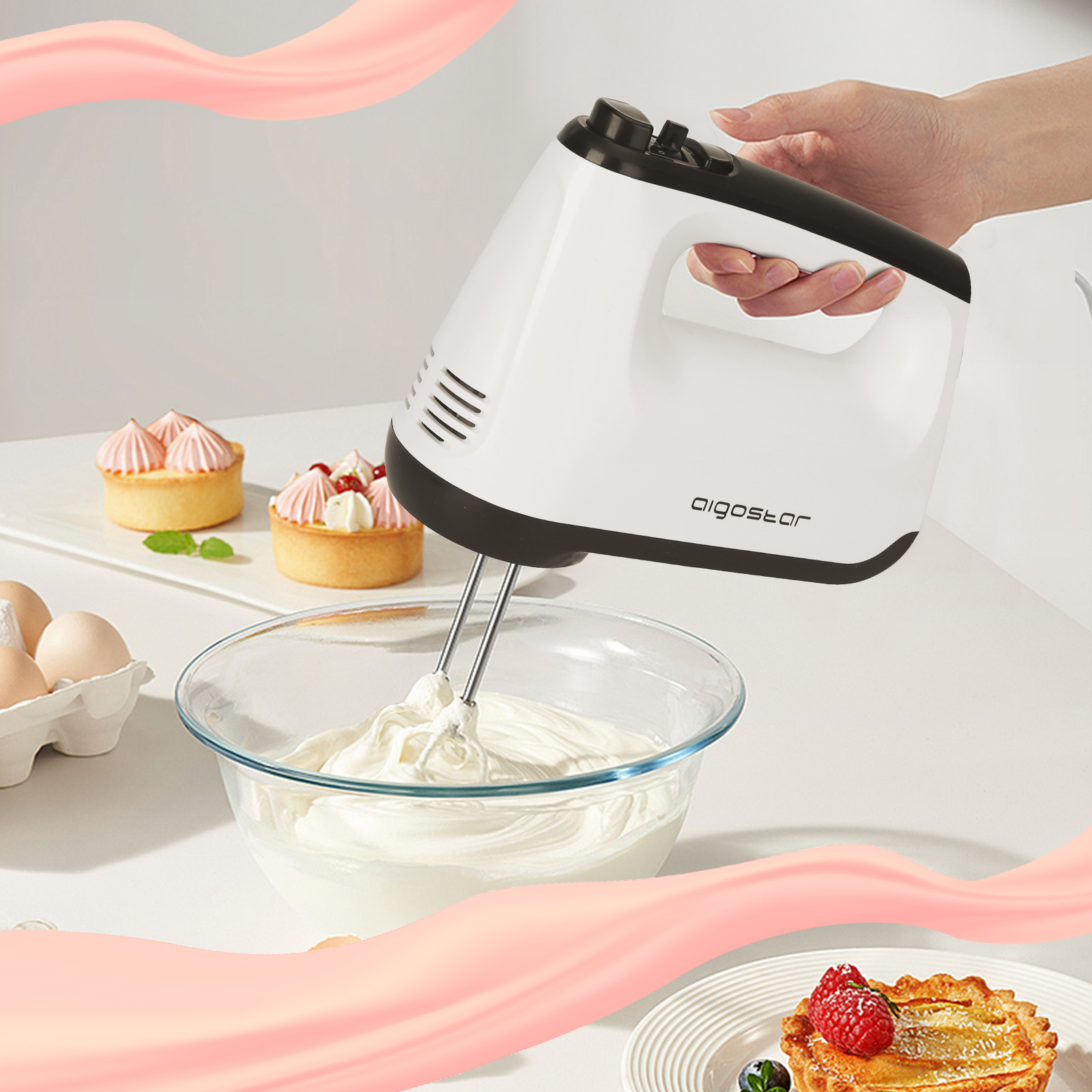 Weißer Handmixer verrührt Sahne in einer Glasschüssel. Kuchen und Eier im Hintergrund.