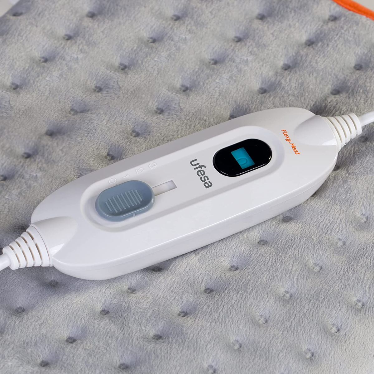 Biały kontroler poduszki grzewczej Ufesa z pokrętłem i wyświetlaczem na szarej tkaninie.