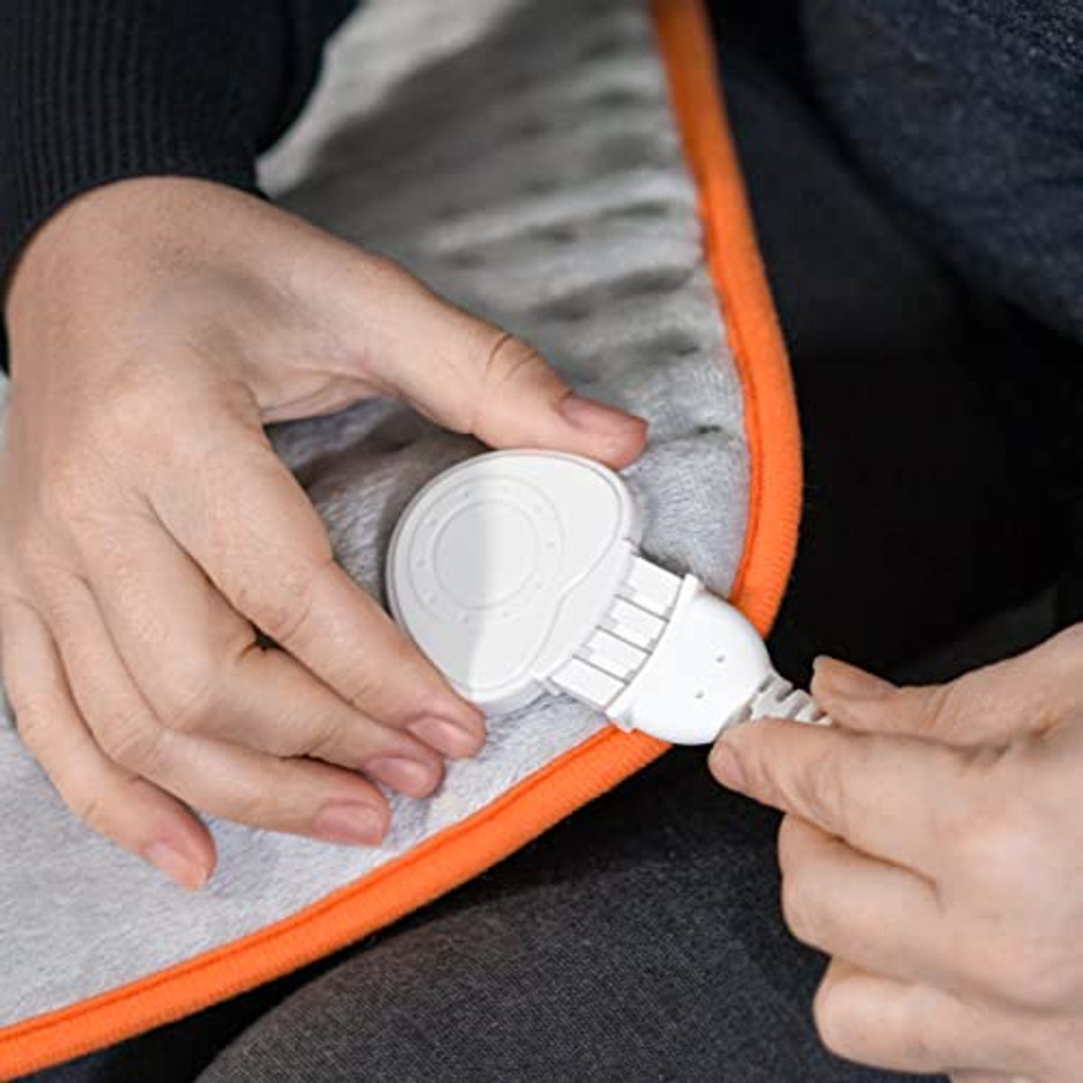 Dłonie trzymają białą wtyczkę poduszki grzewczej. Poduszka ma pomarańczową obwódkę i szary środek.
