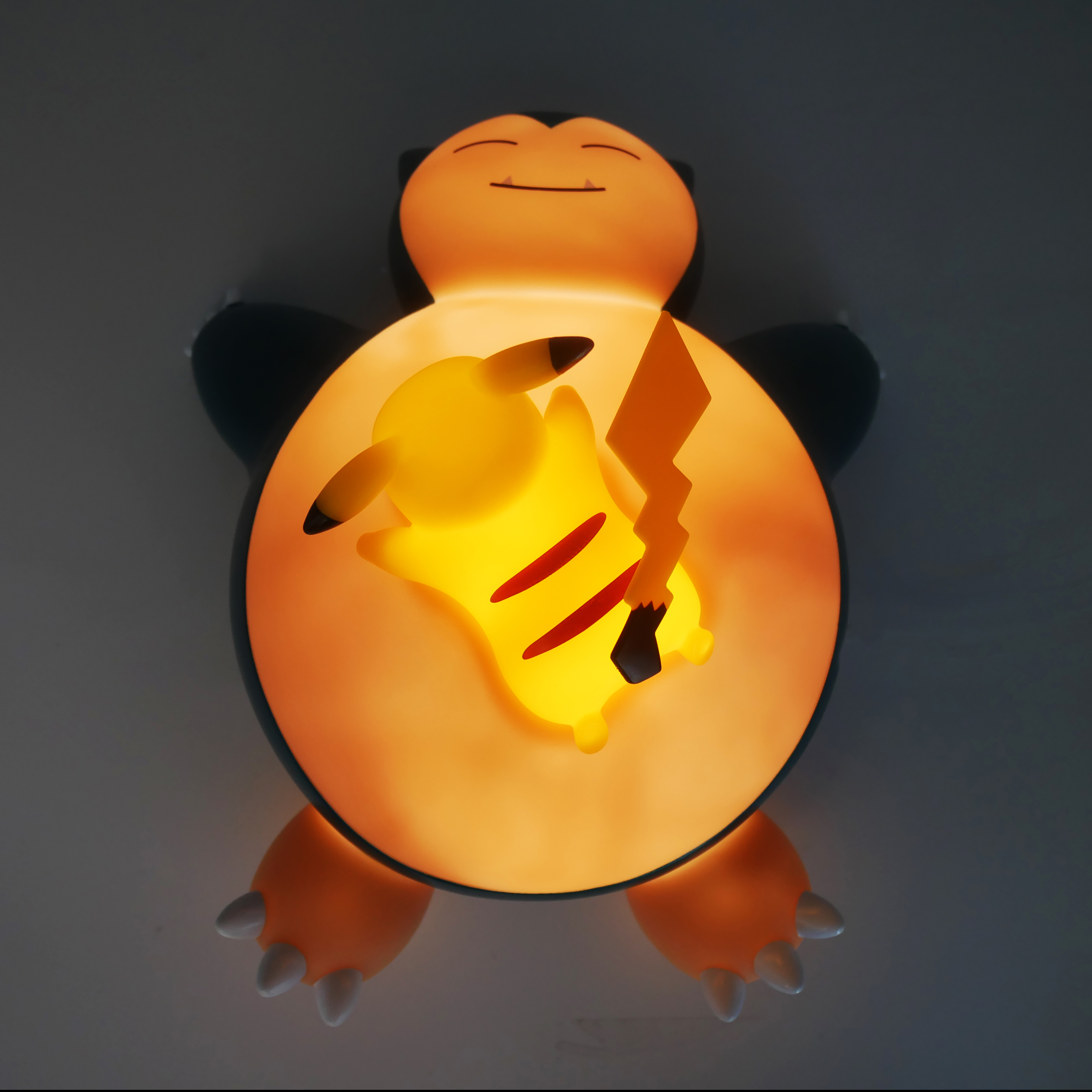 Lampada Pikachu all'interno di una lampada Snorlax. Pikachu è giallo. Snorlax è arancione e blu. Lo sfondo è grigio.