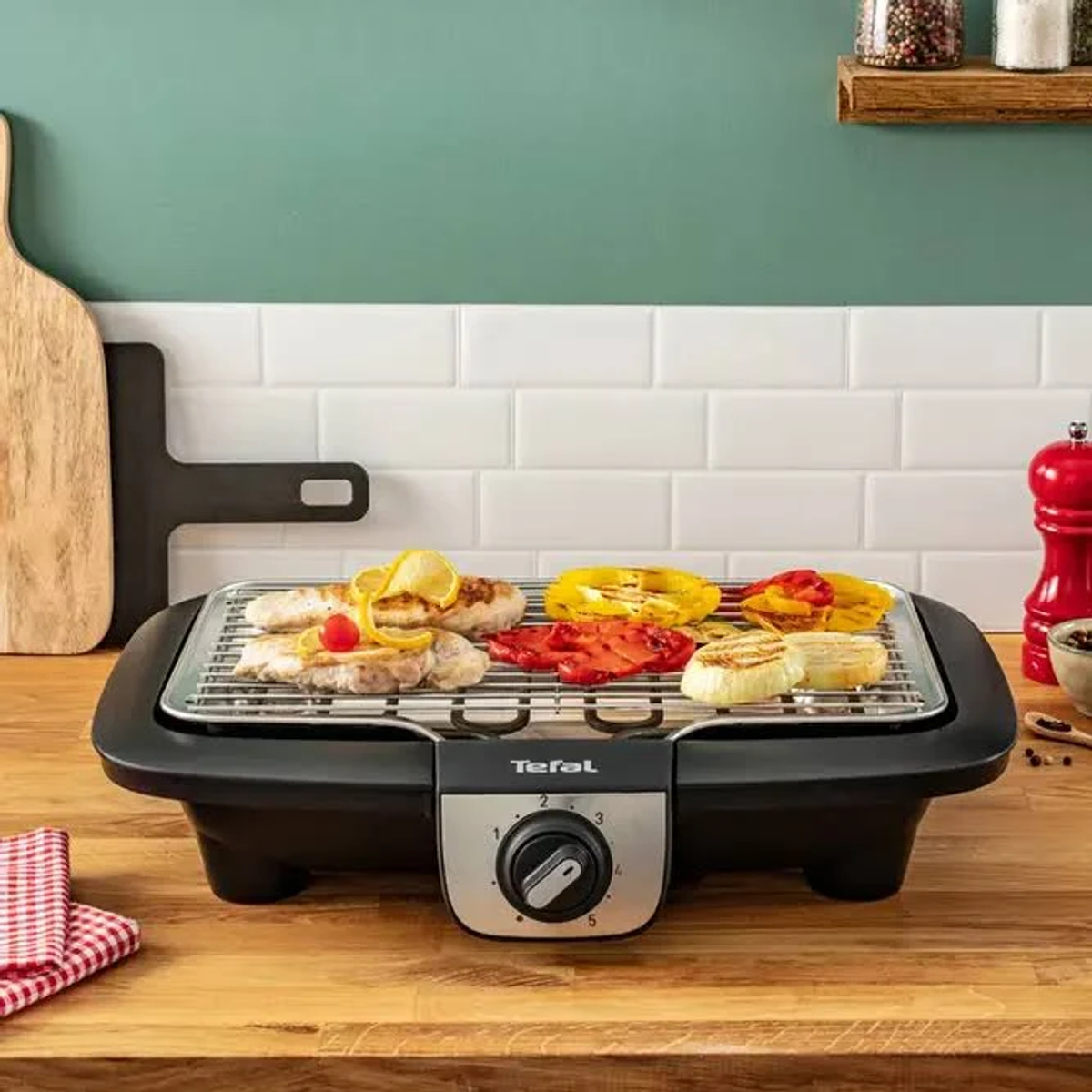 Griglia elettrica Tefal con cibo: peperoni, cipolle e pollo. Tavolo in legno, sfondo di parete piastrellata bianca.