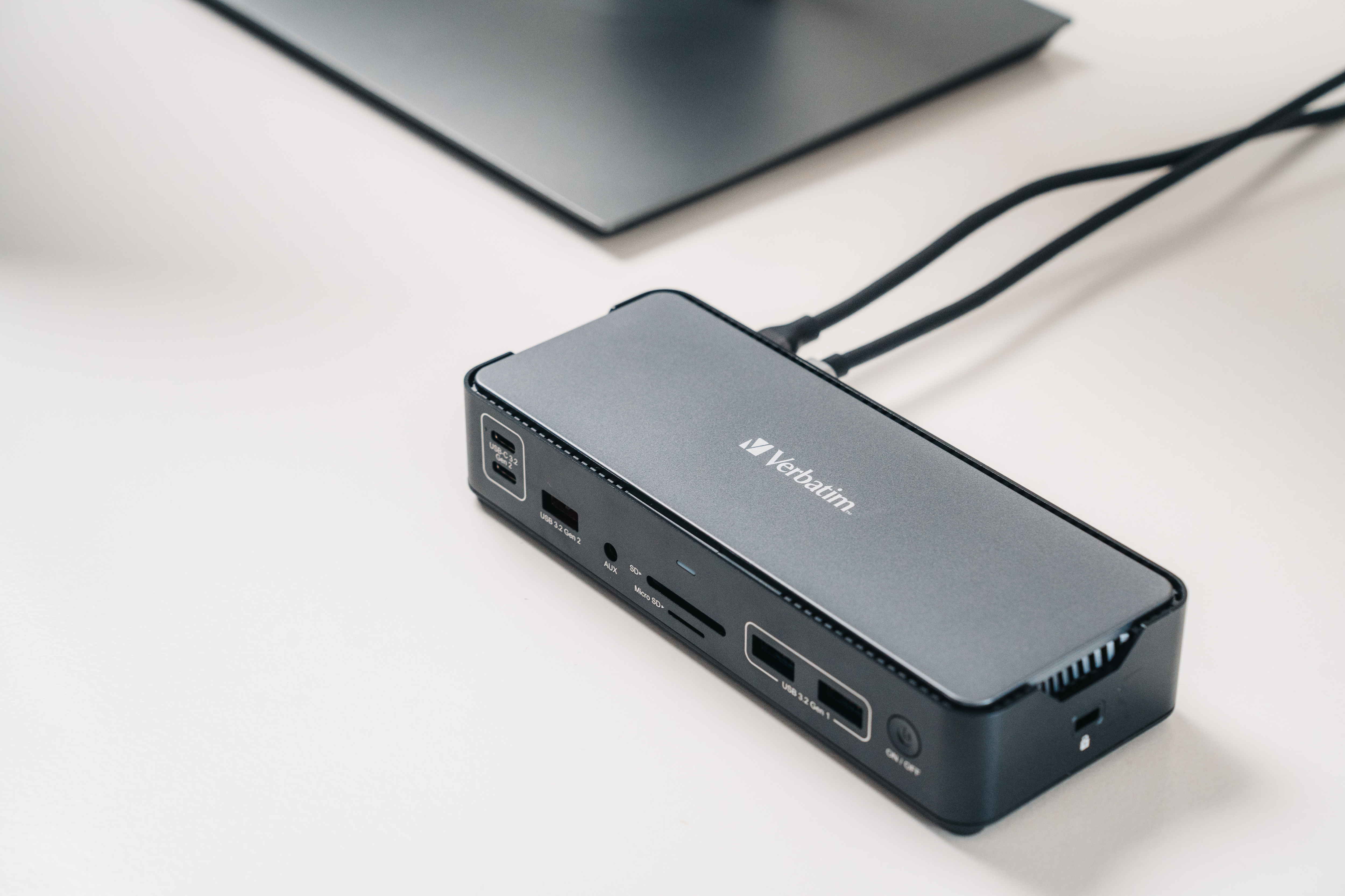 Ein grauer Verbatim-Hub mit Anschlüssen liegt auf einer weißen Oberfläche in der Nähe eines Monitors. Schwarze Kabel.