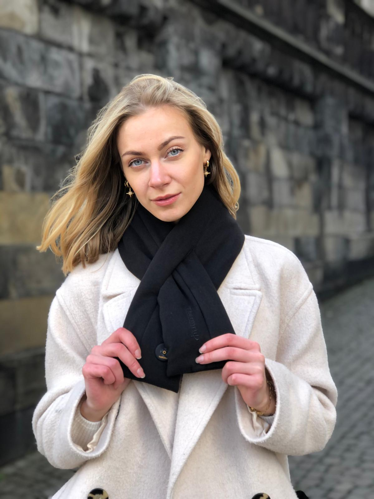 Blonde Frau mit schwarzem Schal und weißem Mantel, stehend vor einer Steinmauer.