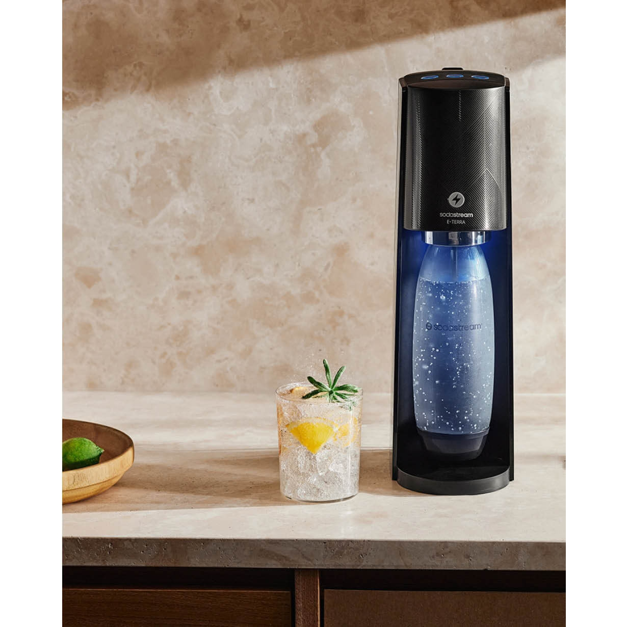 SodaStream Terra Soda Maker mit einem Glas Eis, Zitrone und Rosmarin, vor neutralem Hintergrund.