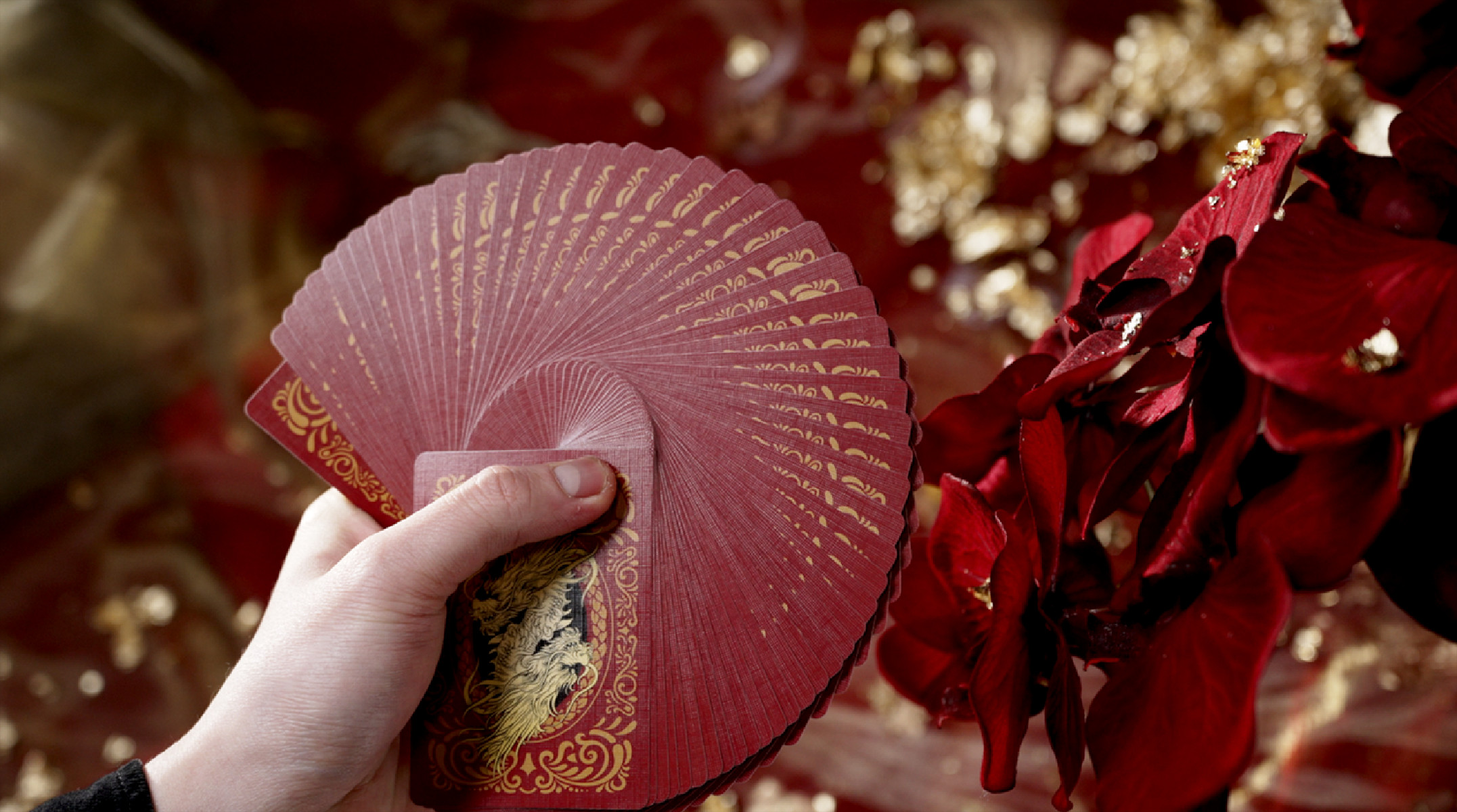 Hand hält gefächerte rote Spielkarten. Goldakzente. Roter Blumengrund.