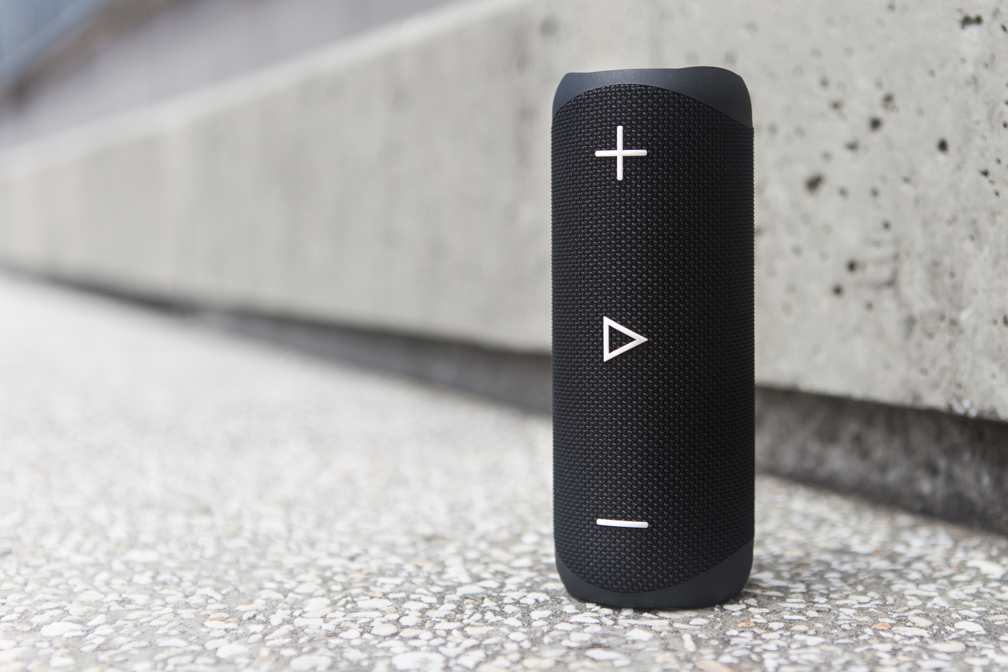 Czarny głośnik przenośny z przyciskami głośności na betonowym tle.