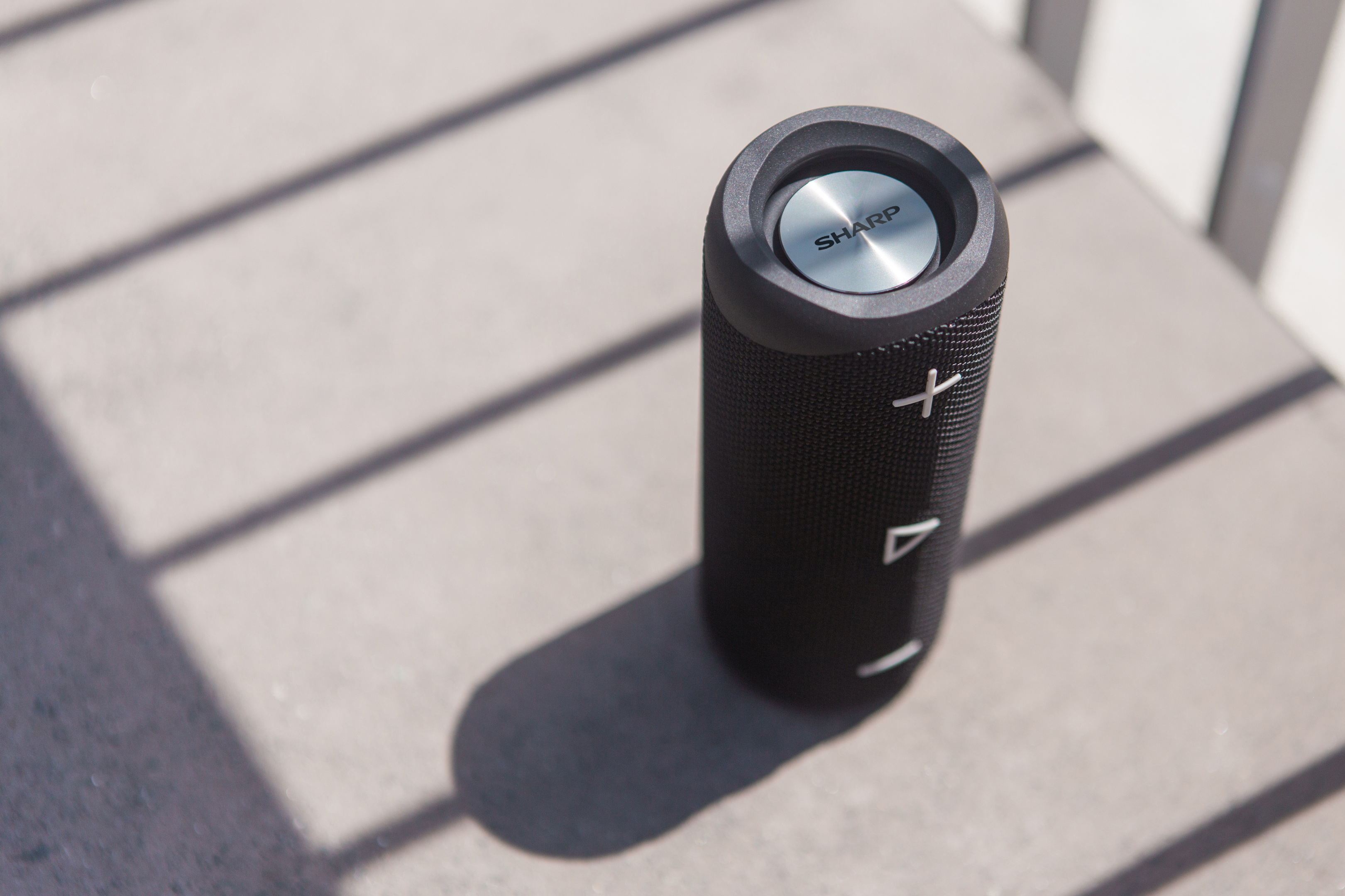 Czarny głośnik Sharp. Cylindryczny kształt, z przyciskami. Cień na szarej powierzchni.