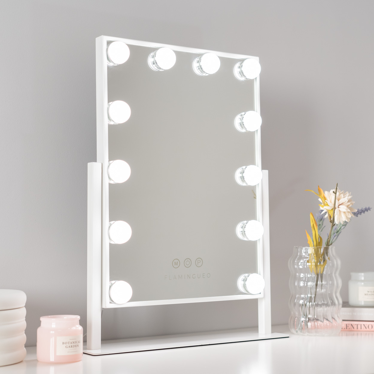 Espejo de tocador blanco con luces, logo FLAMINGUEO y flores en superficie blanca.