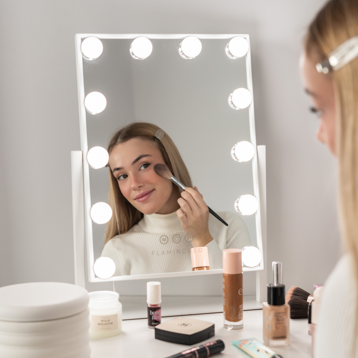 Una mujer maquillándose frente a un espejo de tocador con luces, varios cosméticos en la mesa.