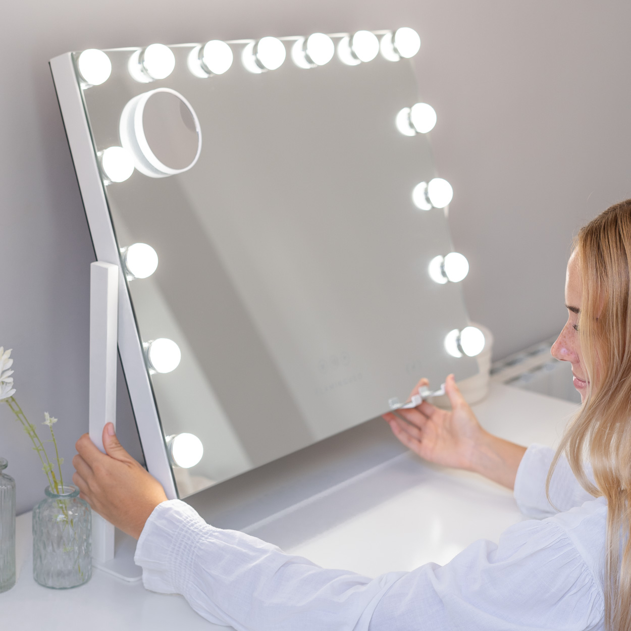 Mujer ajustando un espejo de tocador blanco con luces circundantes.