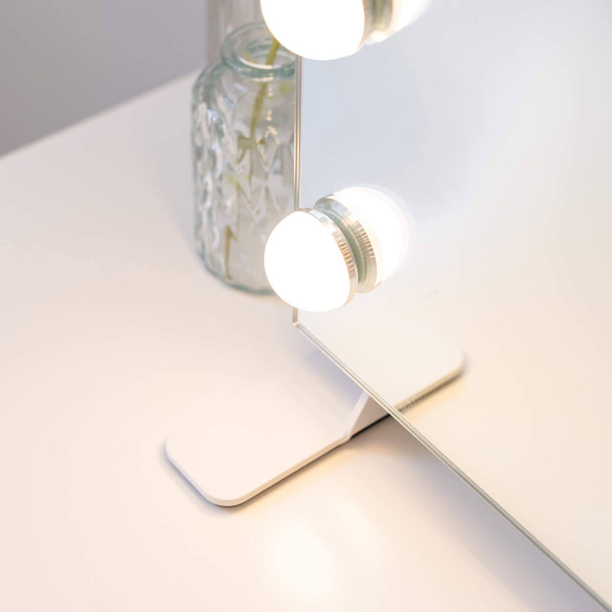 Espejo de tocador blanco con luces y un jarrón de cristal. El espejo está sobre una superficie blanca con dos bombillas.