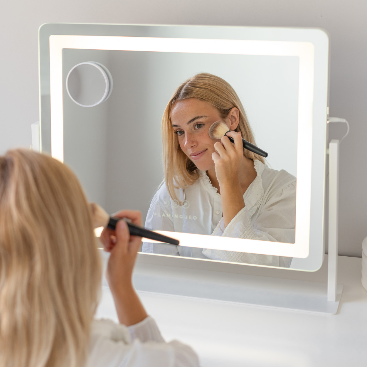 Mujer aplicándose maquillaje frente a un espejo iluminado. Sostiene un pincel, reflejado en el espejo.